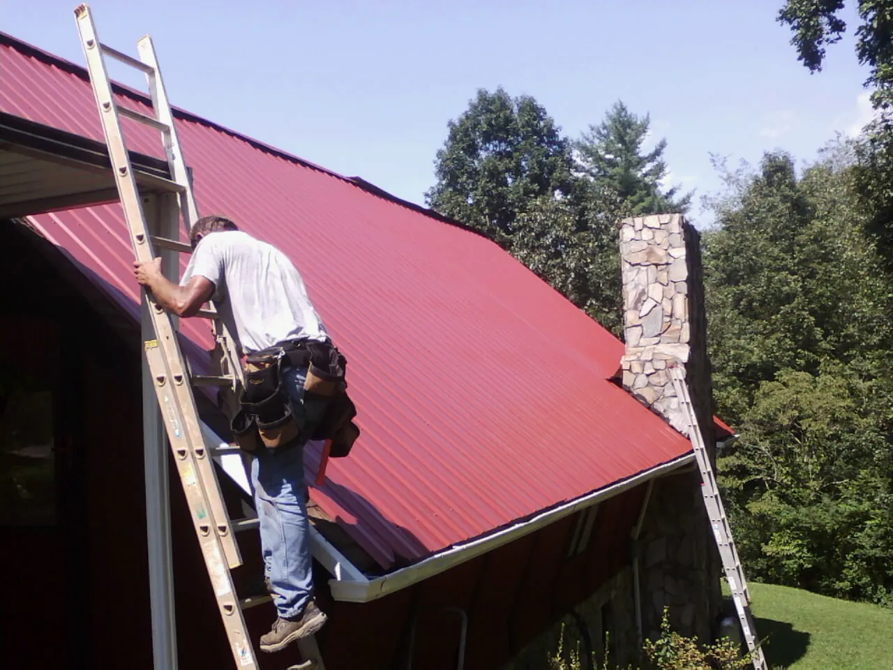 Roofer on a ladder beside a red metal roof and stone chimney — Soffit & Fascia Repair by Roofing Brothers in Oysterville